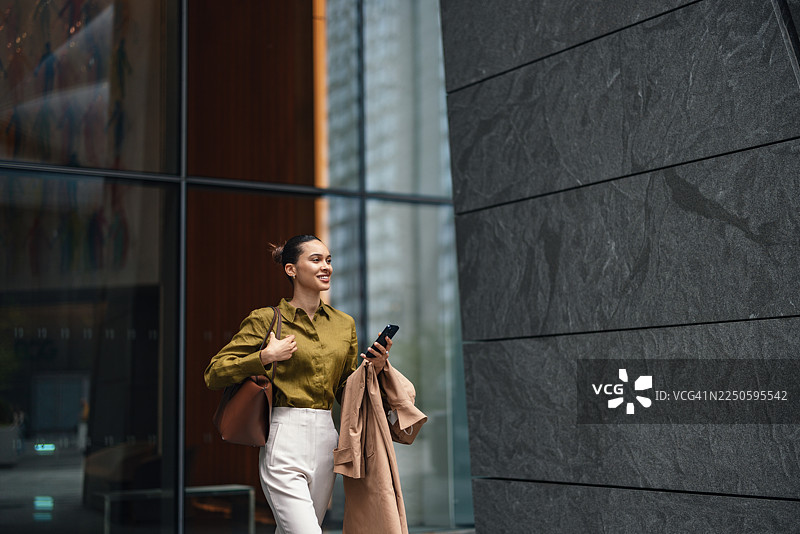 自信的商界女性在现代都市中，身穿外套，手持电话，正在户外行走图片素材