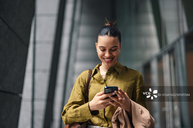 一位穿着商务休闲装的女性在城市街道上微笑着使用智能手机图片素材