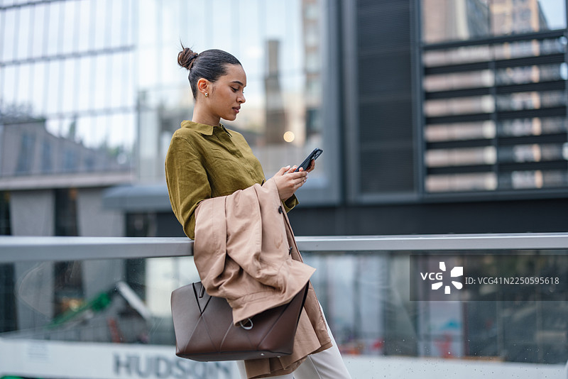 时尚女商人在繁忙的城市街道上查看手机，外套搭在胳膊上图片素材