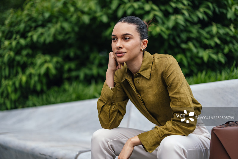 身穿橄榄绿衬衫和米色裤子的商务女性，在户外长椅上若有所思图片素材