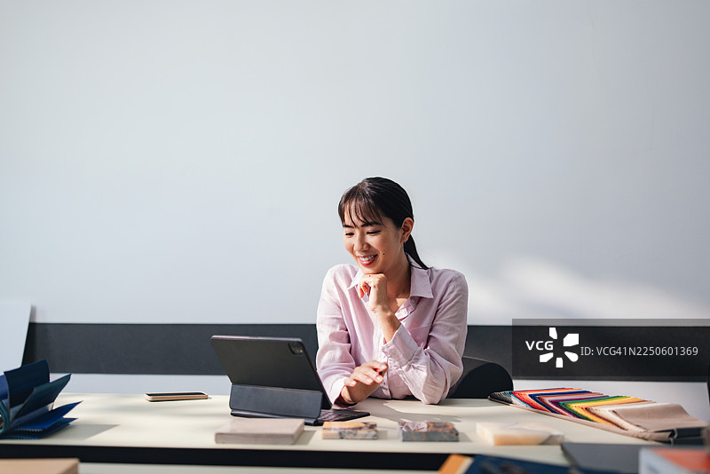 现代办公室里，一位女商人在书桌旁使用平板电脑，周围散布着彩色的样品和文件。图片素材