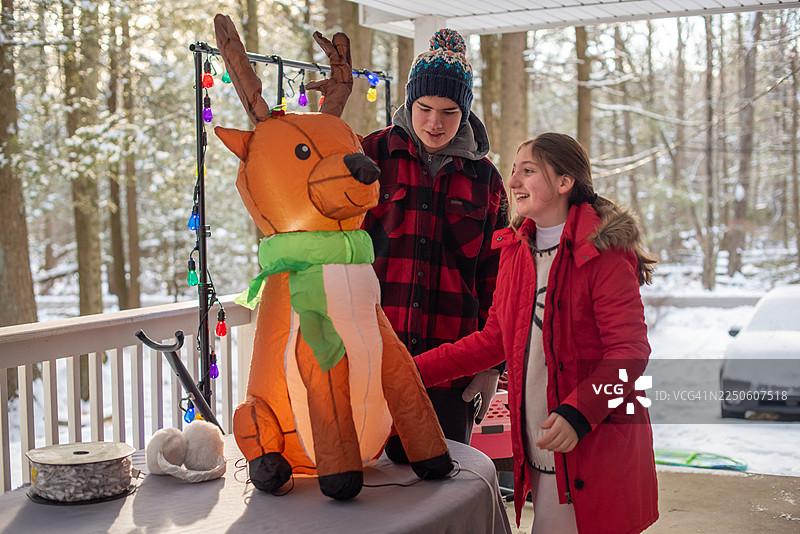 快乐的兄弟姐妹在冬日假期里，在覆盖着雪的露台上装饰充气驯鹿。小女孩和患有自闭症的年轻男子在山间小屋布置圣诞装饰。图片素材