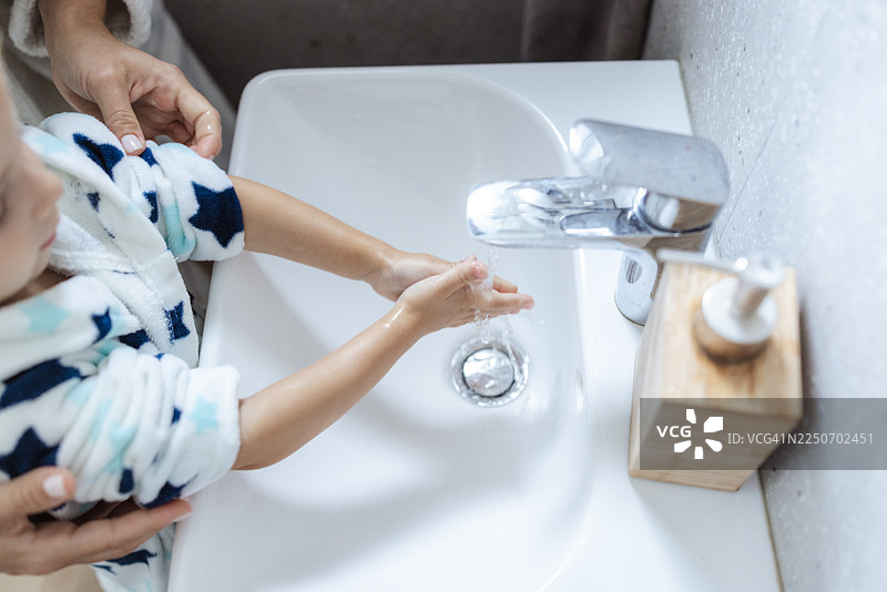 妈妈在浴室的水槽边帮助蹒跚学步的孩子洗手，促进卫生和亲子关系图片素材