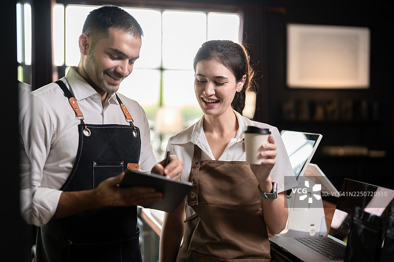 两位穿着围裙的咖啡师，手持平板电脑和咖啡杯，开心地讨论着工作，可能是在咖啡馆里管理订单或安排日程。图片素材