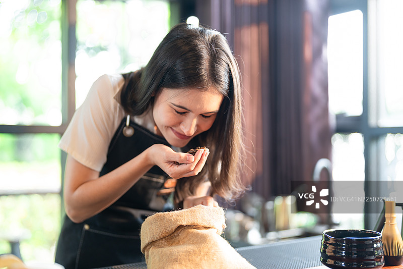 一位穿着围裙的女士在一个光线充足的房间里，微笑着检查一个粗麻布袋里的东西，这个地方可能是一家咖啡馆或商店。图片素材