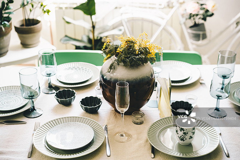 一张布置得明亮而时尚的餐桌，上面摆放着陶瓷盘子、玻璃杯和装饰碗，中心是一个装满黄色鲜花的大花瓶。图片素材