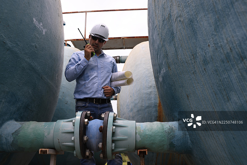 一位戴着安全帽和太阳镜的建筑专业人士，在大型水箱和阀门工地使用对讲机和蓝图进行工作，强调工业环境中的沟通与团队合作图片素材