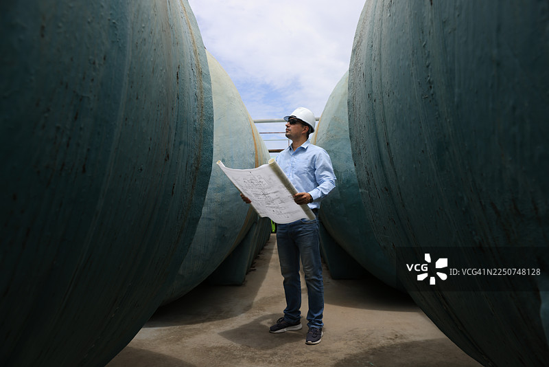 一位戴着安全帽和太阳镜的建筑专业人士或高级工程师，站在大型水箱之间，正在查看蓝图。图片素材