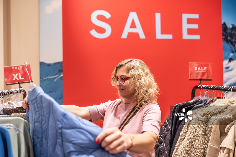一位女士在黑色星期五期间在一家商店里看着一件蓝色的绗缝夹克。背景中有一个醒目的红色“促销”标志，表明她正在寻找便宜货。图片素材