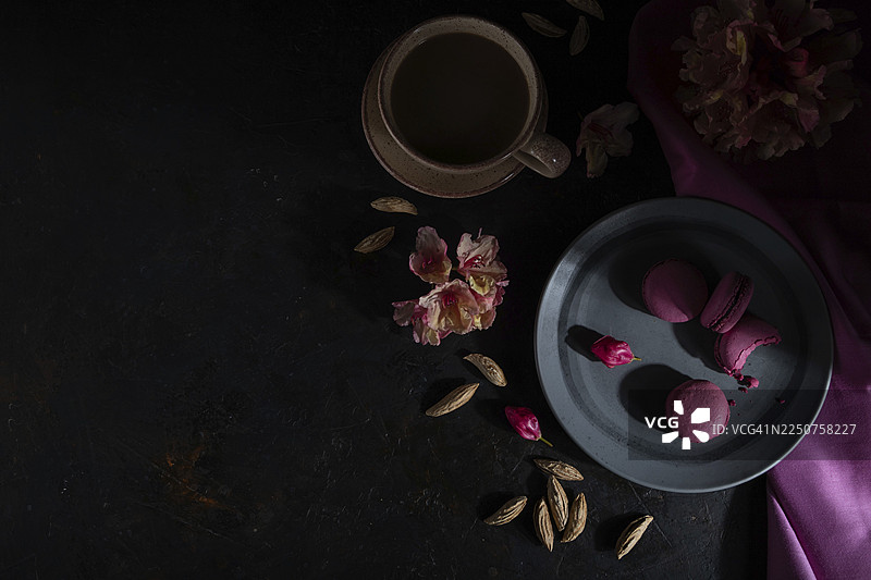 黑色的混凝土背景和粉红色的纺织品上放着紫色的马卡龙或马卡龙蛋糕，旁边还有一杯咖啡。硬光，低调。俯视，平面图，留白。图片素材