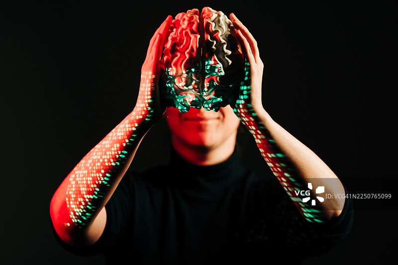 一个人在黑暗的背景下，手持被数字代码投影照亮的脑模型。图片素材