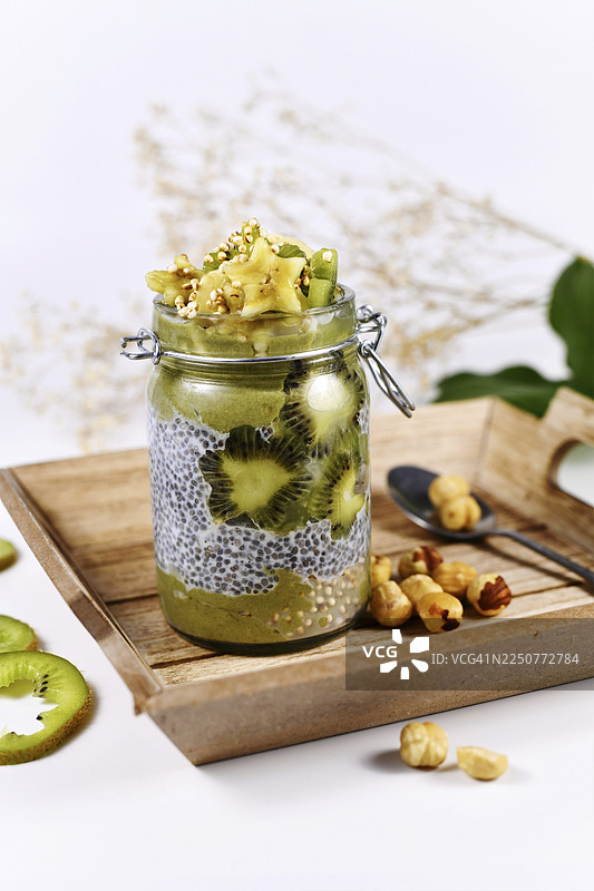 木质托盘上绿色的水果冰沙分层装在玻璃杯中，上面是奇亚籽布丁，点缀着星形香蕉和猕猴桃片。图片素材