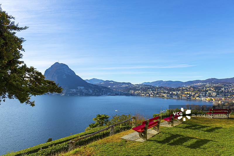 瑞士卢加诺提契诺卡斯塔尼奥拉圣米歇尔公园的长凳、树枝、卢加诺湖、城市和远处的山以及蓝天图片素材