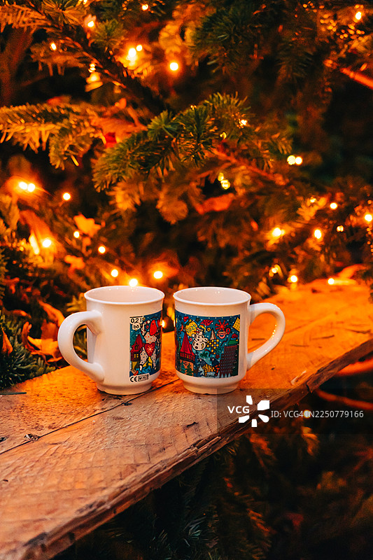 舒适的圣诞场景，木桌上放着两个节日马克杯，旁边是闪烁的圣诞树灯光图片素材