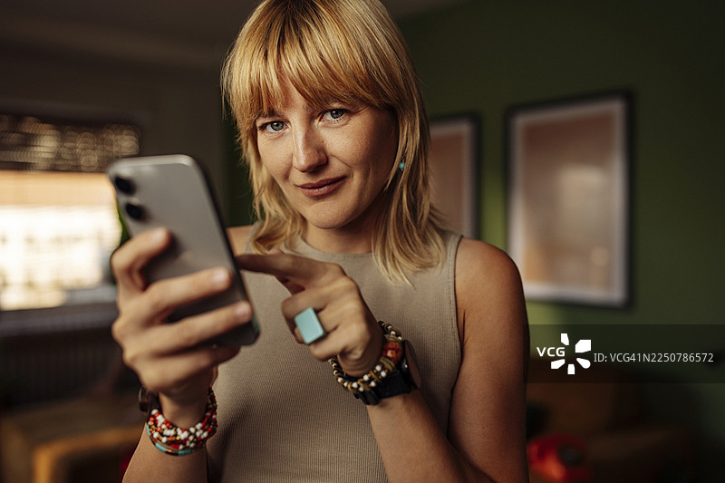 一位女士在室内使用智能手机，手腕上戴着手镯，脸上带着微笑图片素材