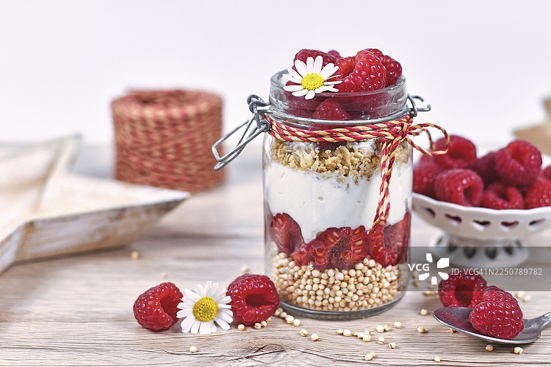 健康的覆盆子水果甜点，用斯凯尔酸奶、格兰诺拉麦片和膨化藜麦谷物分层装在罐子里，周围摆放着配料。图片素材