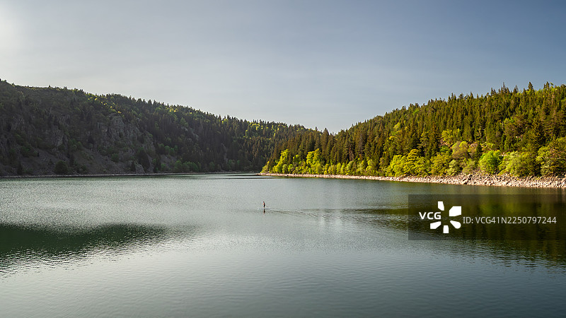 春季白天从湖岸看到的白湖（Lac Blanc）全景，这是孚日山脉（Vosges mountains）著名的自然地标，树木倒映在水中，一位无法辨认的男子正在桨板上划行，地点位于法国阿尔萨斯上莱茵省奥尔贝（Orbey - Haut-Rhin - Alsace - France）。图片素材