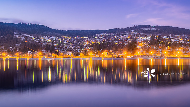 冬季，从湖岸看到的法国东部洛林孚日山脉热拉尔梅湖（Lac de Gérardmer）平静的水面倒映着被雪覆盖并亮灯的房屋和木屋的美景，热拉尔梅图片素材