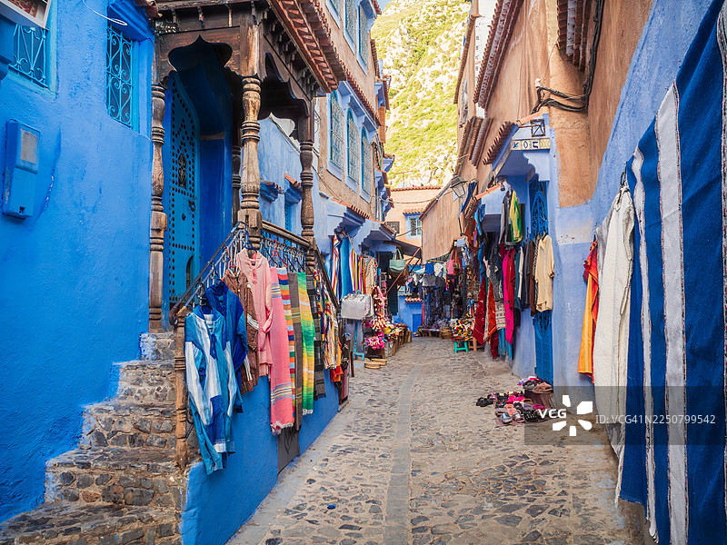 摩洛哥丹吉尔-得土安-胡塞马地区舍夫沙万“蓝色之城”麦地那中心的鹅卵石购物小巷，街道两旁是售卖服装和纺织品的纪念品和礼品店，商品陈列在街上。图片素材