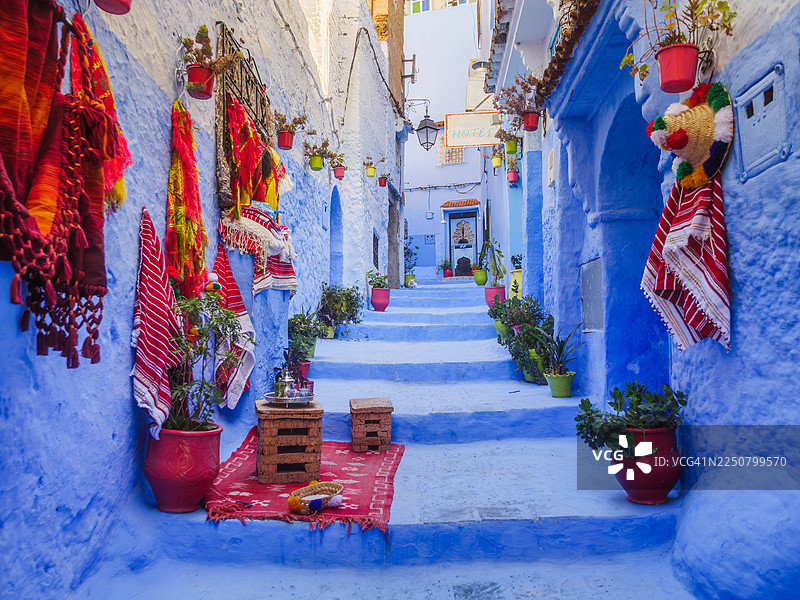 摩洛哥丹吉尔-德托万-胡塞马地区舍夫沙万麦地那的向上巷道,有楼梯,两旁是蓝色的房屋,装饰着盆栽植物,墙上挂着帽子和披肩,还有一套用于摆拍游客的茶具,这里是著名的“蓝色之城”。图片素材
