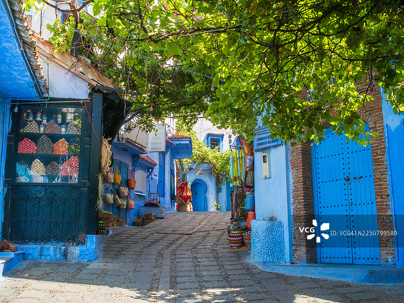 摩洛哥丹吉尔-得土安-胡塞马地区，舍夫沙万这座著名的“蓝色之城”麦地那（老城区）内，一条铺满鹅卵石的向上街道，街道两旁是售卖服装、香皂、包袋和篮子等纪念品的商店，商品陈列在街边，街道被一棵大树覆盖。图片素材