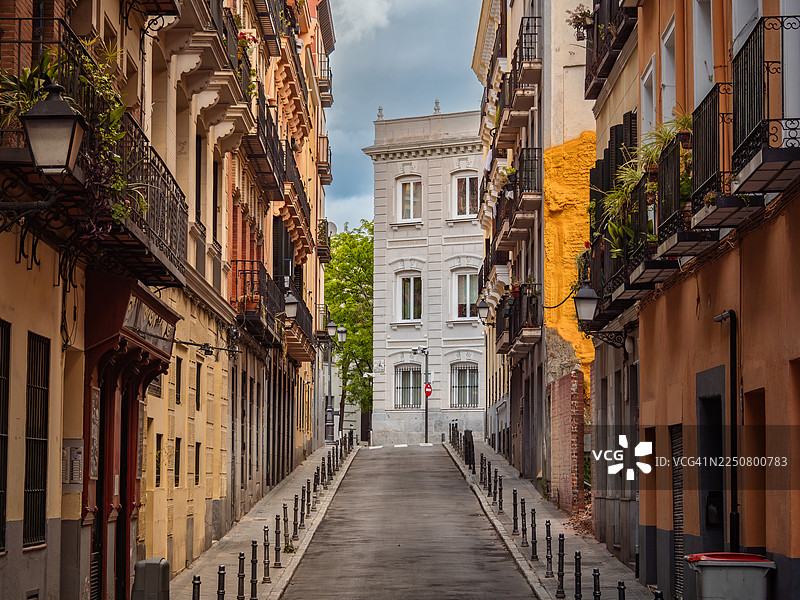 马德里自治区马德里市拉拉蒂纳区一条狭窄的上坡街道的近景，只有一条车道和人行道，街道两侧是色彩鲜艳的住宅楼和公寓，装饰着锻铁阳台和窗台花箱。图片素材
