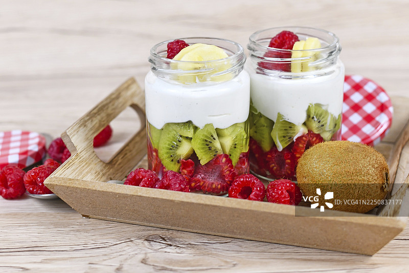 两个装有健康分层零食的杯子，配有新鲜覆盆子、奇异果和酸奶，放在木托盘上，周围摆放着配料图片素材