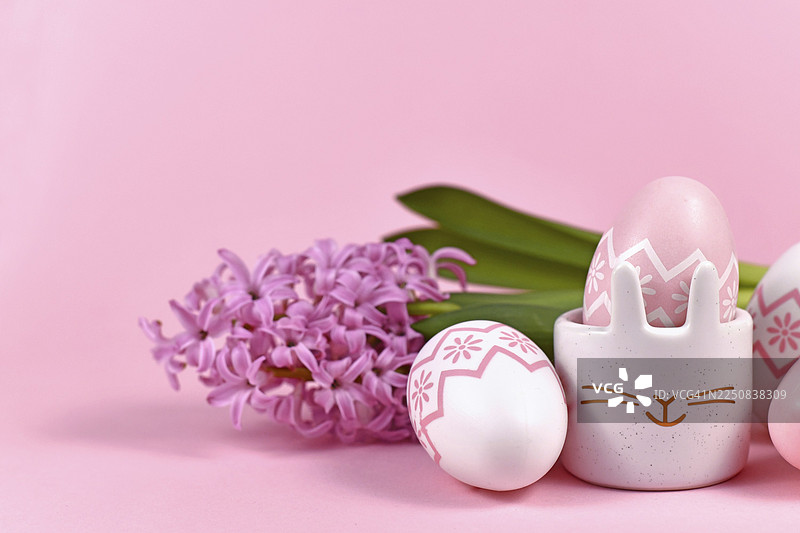 粉白色彩绘的复活节彩蛋和可爱的兔子造型蛋杯，背景为粉色，上面装饰着风信子春花。图片素材