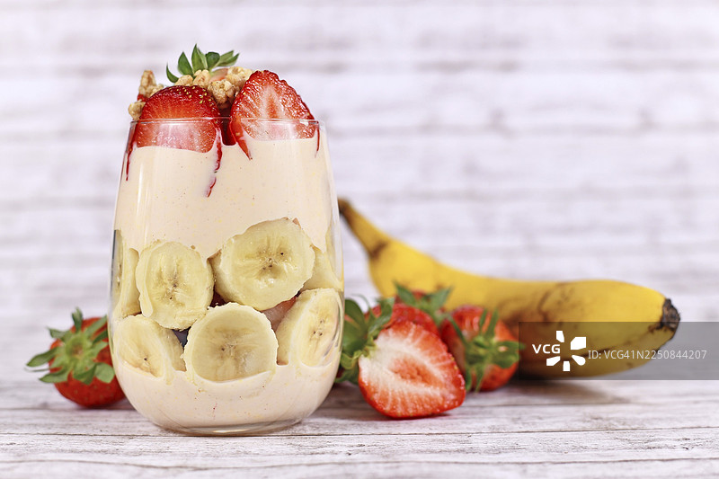 健康的香蕉和草莓水果夏季甜点，搭配酸奶和格兰诺拉麦片，分层装在玻璃杯中，周围环绕着新鲜食材。图片素材