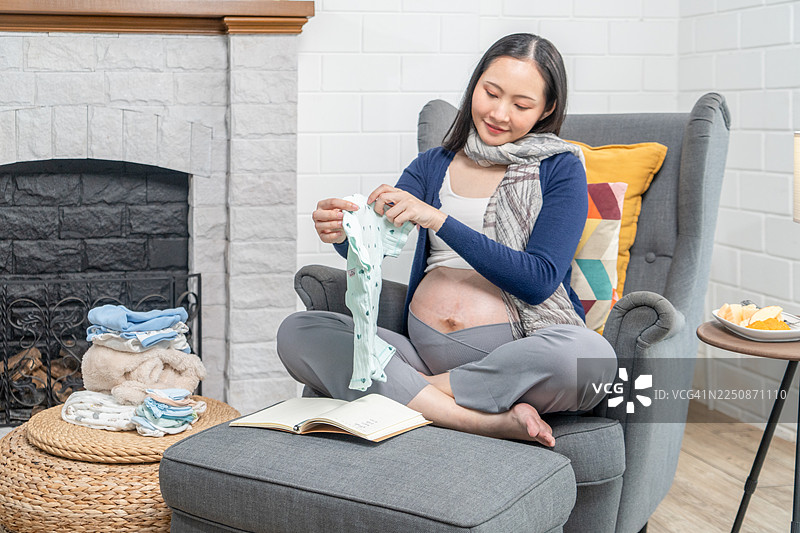 一位怀孕的亚洲单身母亲即将分娩。为迎接新生儿准备衣物。一位年轻女子在床上将女儿的衣服叠好放进篮子里，她对即将到来的宝宝感到欣喜。图片素材