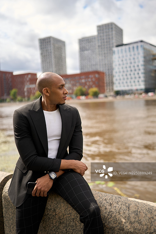 穿着西装外套的男士坐在河边的石墙上，望向远方，背景是现代高楼大厦。图片素材