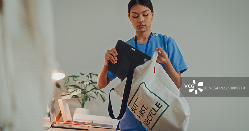 年轻的亚裔女护士穿着蓝色工作服，在卧室里套上一件米色毛衣。护士生活方式概念。图片素材