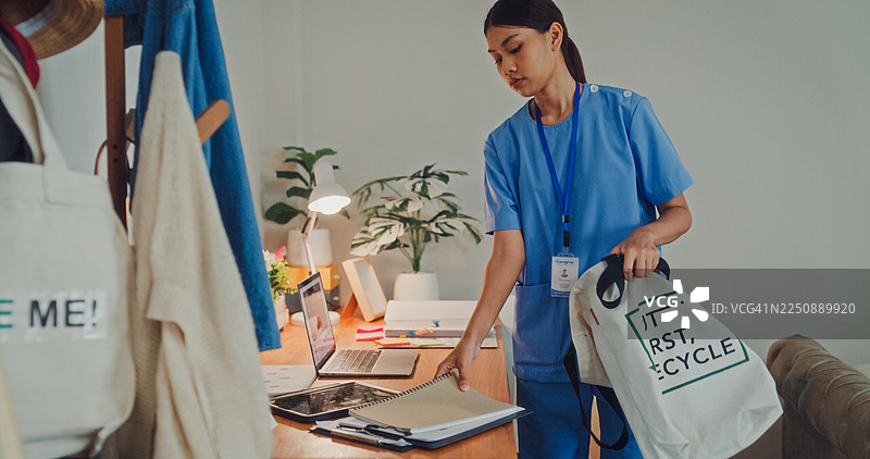 年轻的亚洲女护士穿着蓝色工作服，在卧室里套上一件米色毛衣。护士生活方式概念。图片素材