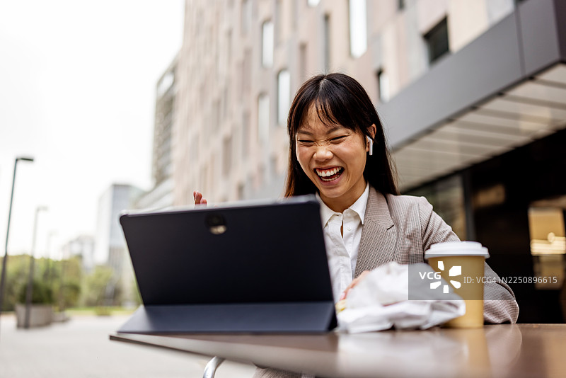 一位女士在户外咖啡馆微笑着使用笔记本电脑，旁边放着咖啡杯和纸袋。图片素材