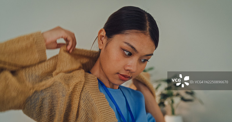 卧室里，一位身穿蓝色工作服的年轻亚洲女护士正在穿米色毛衣。护士生活方式概念。图片素材