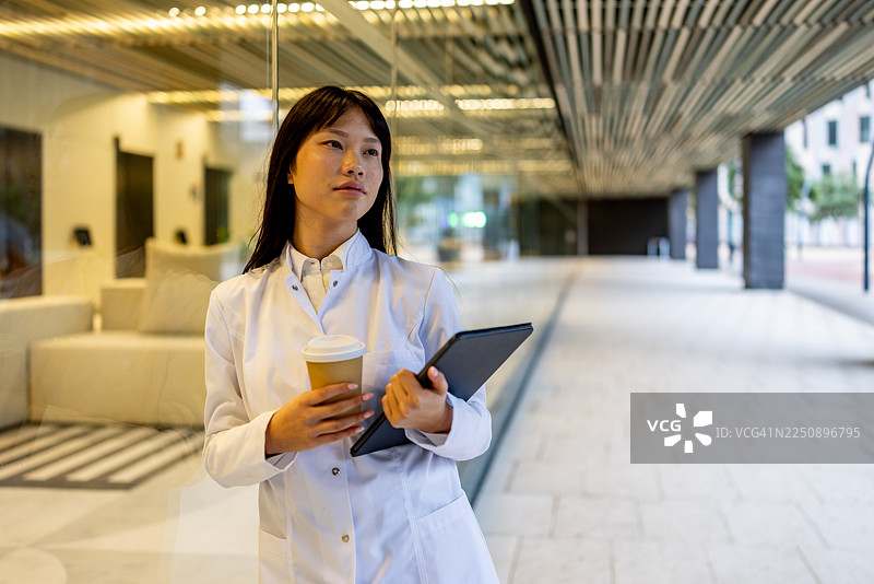 Professional Woman in White Lab Coat Holding Coffee and Tablet in Modern Office Lobby图片素材