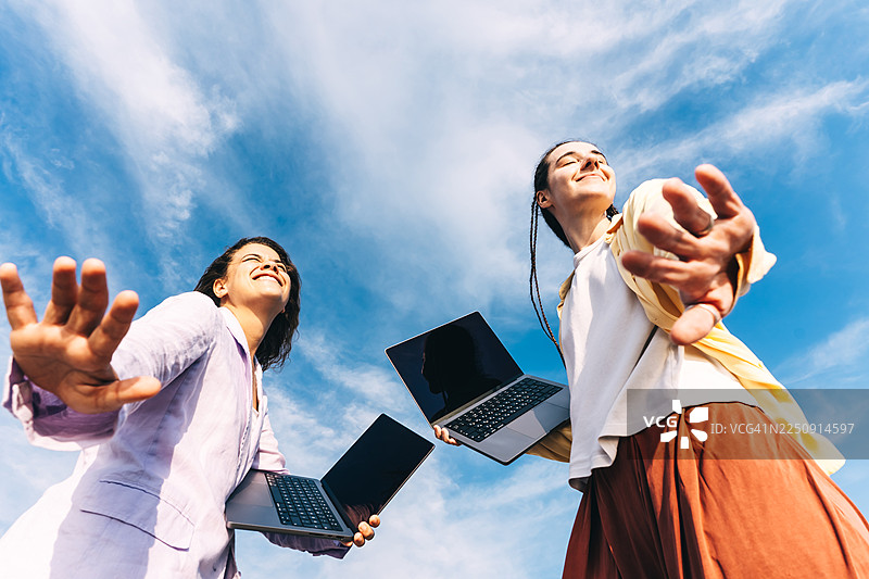 两位年轻女子在生机勃勃的蓝天下户外拿着笔记本电脑，微笑着向前伸出手。图片素材