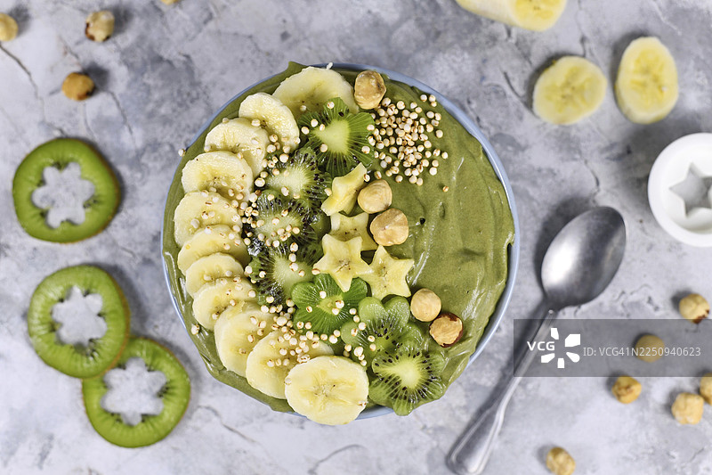 灰色背景上放着绿色羽衣甘蓝水果冰沙碗，上面装饰着星星形状的香蕉、猕猴桃、榛子和膨化藜麦谷物图片素材