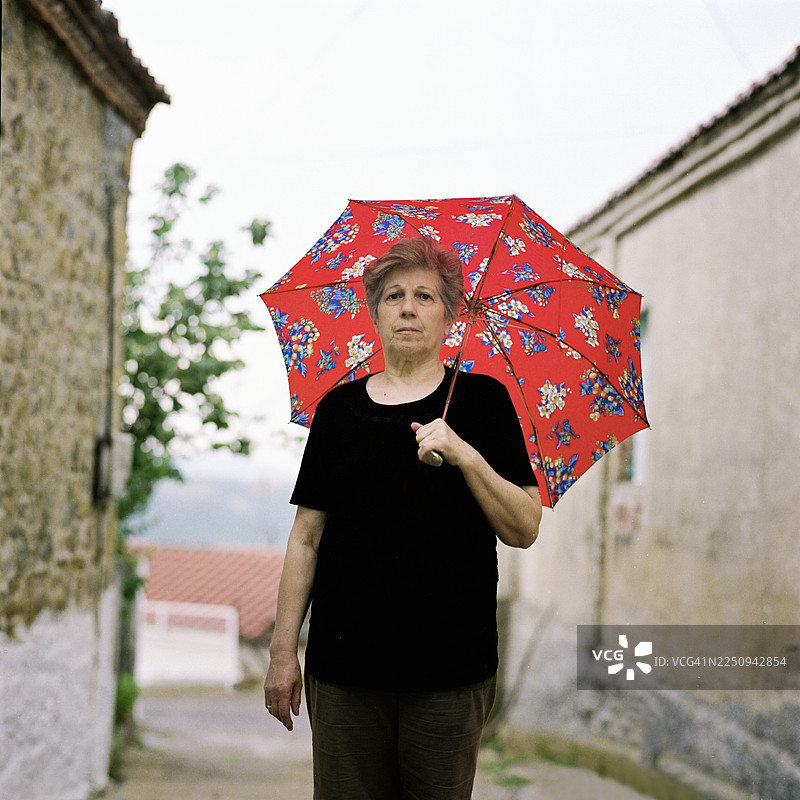 女人站在狭窄的街道上，撑着红伞，身处乡村。图片素材
