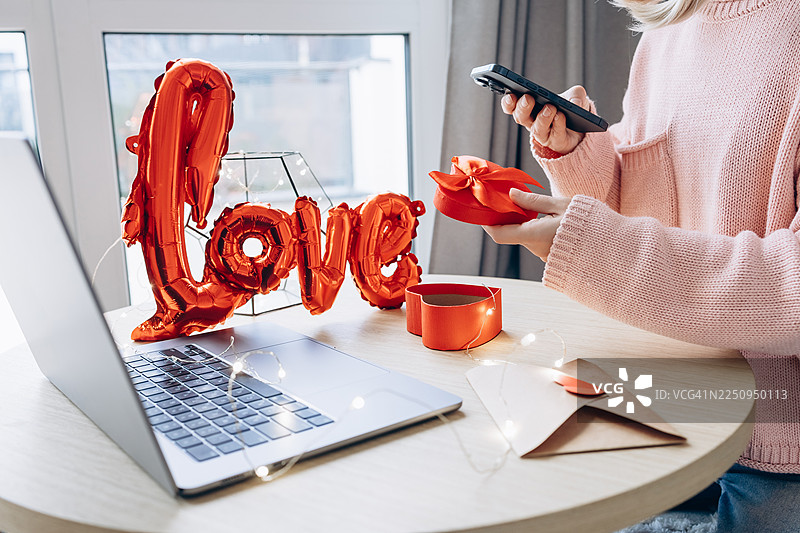 女子在办公桌前使用智能手机，手中拿着红色的心形礼品盒，桌上还有笔记本电脑和“爱”字气球装饰。图片素材