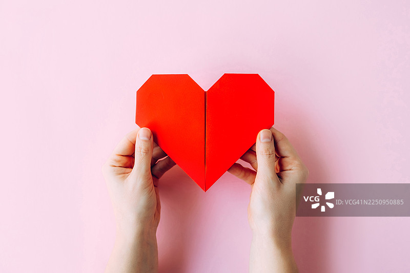 柔粉色背景上两只手捧着一颗红色纸心，有留白区域的特写。图片素材