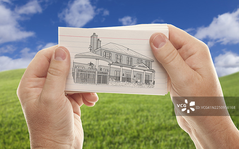 男人的手拿着一叠纸，纸上画着一栋房子，背景是空旷的草地和天空。图片素材