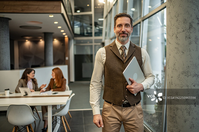 成功的商人，身处现代办公室，手持笔记本电脑，面带微笑图片素材