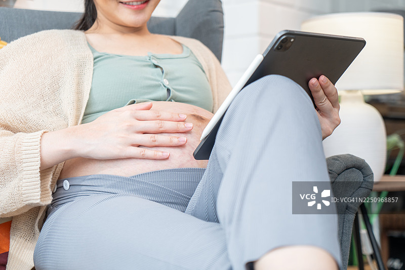年轻孕妇穿着休闲服，挺着肚子，手里拿着平板电脑，上面显示着超声波图像，她坐在地板上，在家或酒店的室内起居室里休息、放松、享受空闲时光。图片素材