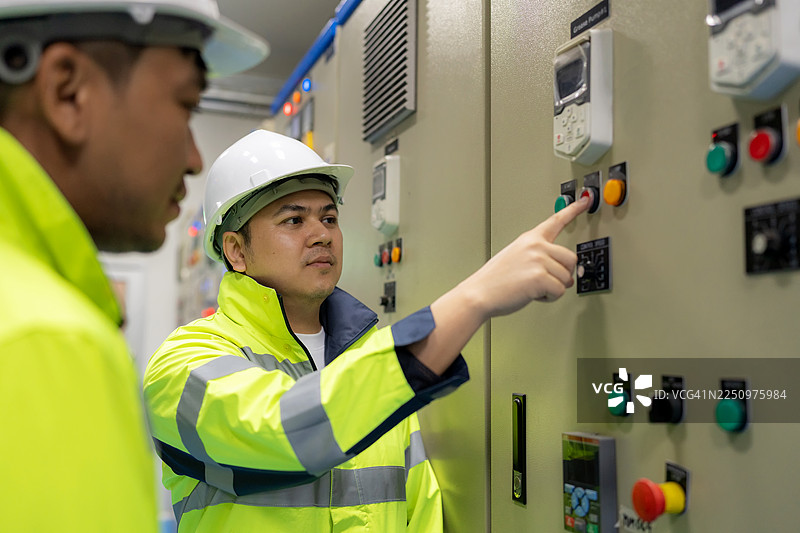 两位电工在MDB控制室操作开关并检查电力流，他们在一块数字平板上记录数据，周围是开关设备控制面板、配电盘、电缆、电路板、保护熔断器盒、高压标志和技术。图片素材