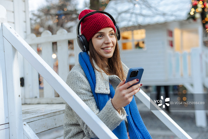一位身着红色针织帽和蓝色围巾的女士在冬天户外用智能手机听音乐图片素材