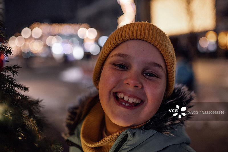 戴着芥末黄色绒线帽、身穿冬外套的快乐小女孩在节日夜晚户外玩耍图片素材