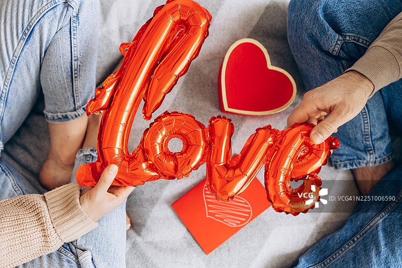 俯视视角，两个人坐在一起，盖着柔软的毯子，手中拿着一个闪亮的红色“爱心”铝箔气球。图片素材