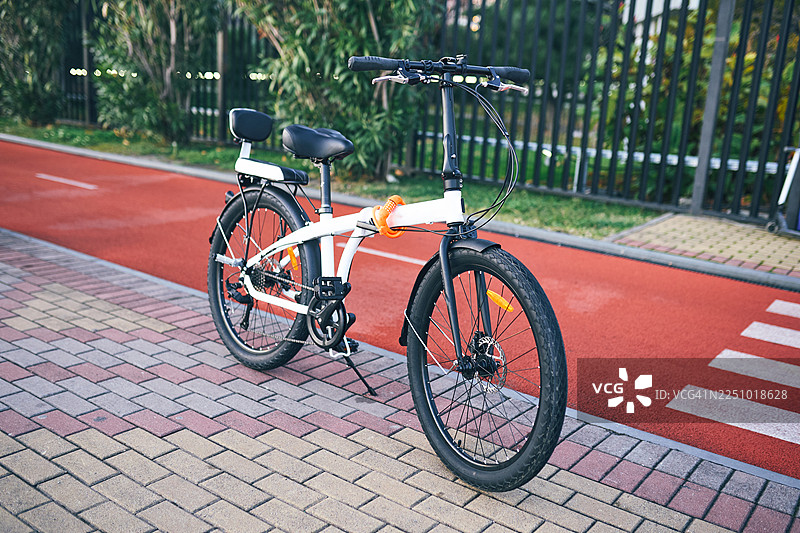 城市公园里，白色自行车停在红色的自行车道上，周围有绿植和栅栏。图片素材