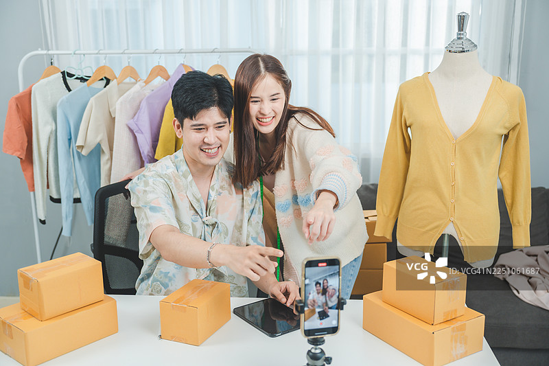 年轻的企业家们正在直播服装业务，展示时尚的服装，周围堆满了包裹，以提高在线零售的可见度。图片素材